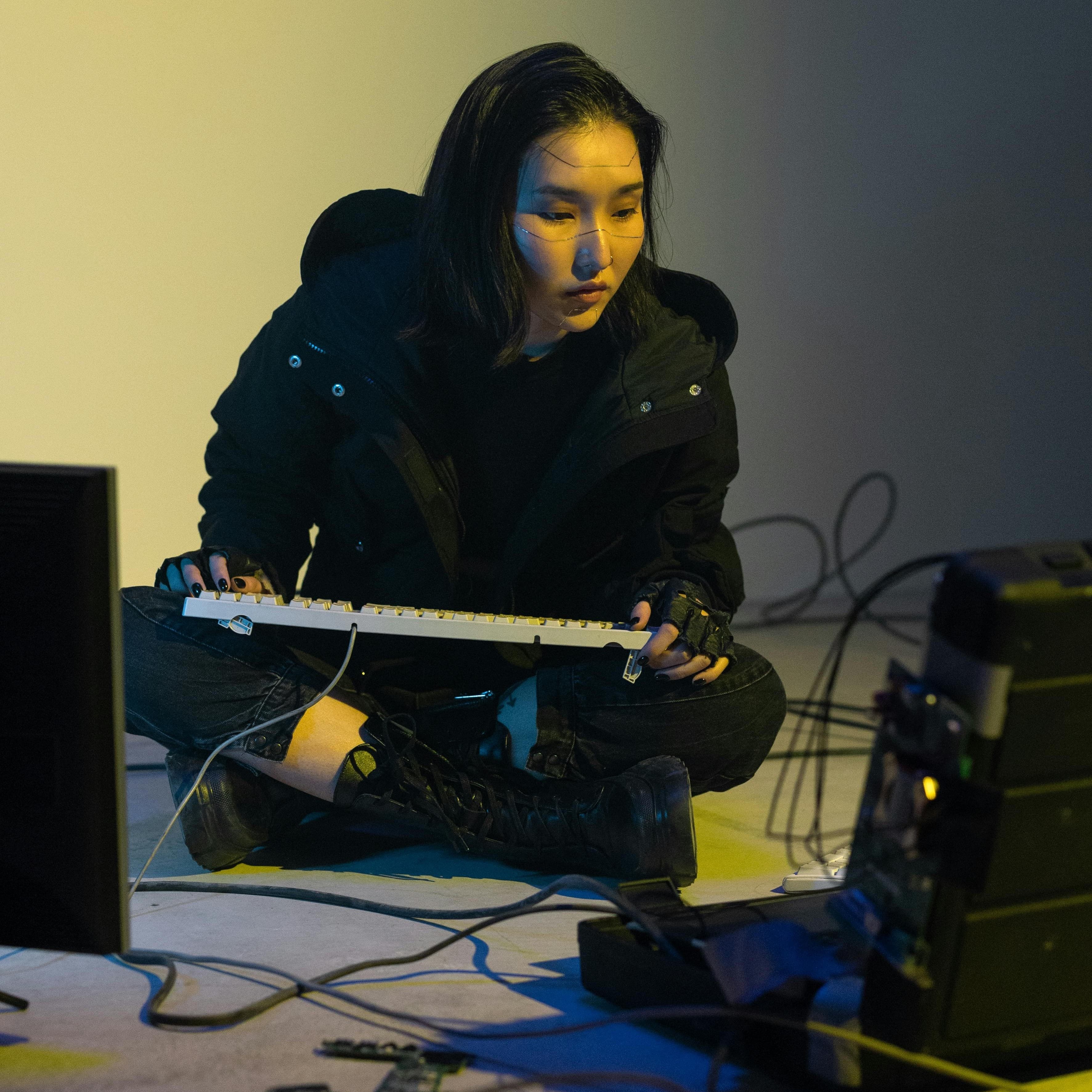 Person with cultural face tattoos sitting on the floor with a keyboard in their lap, viewing a computer.