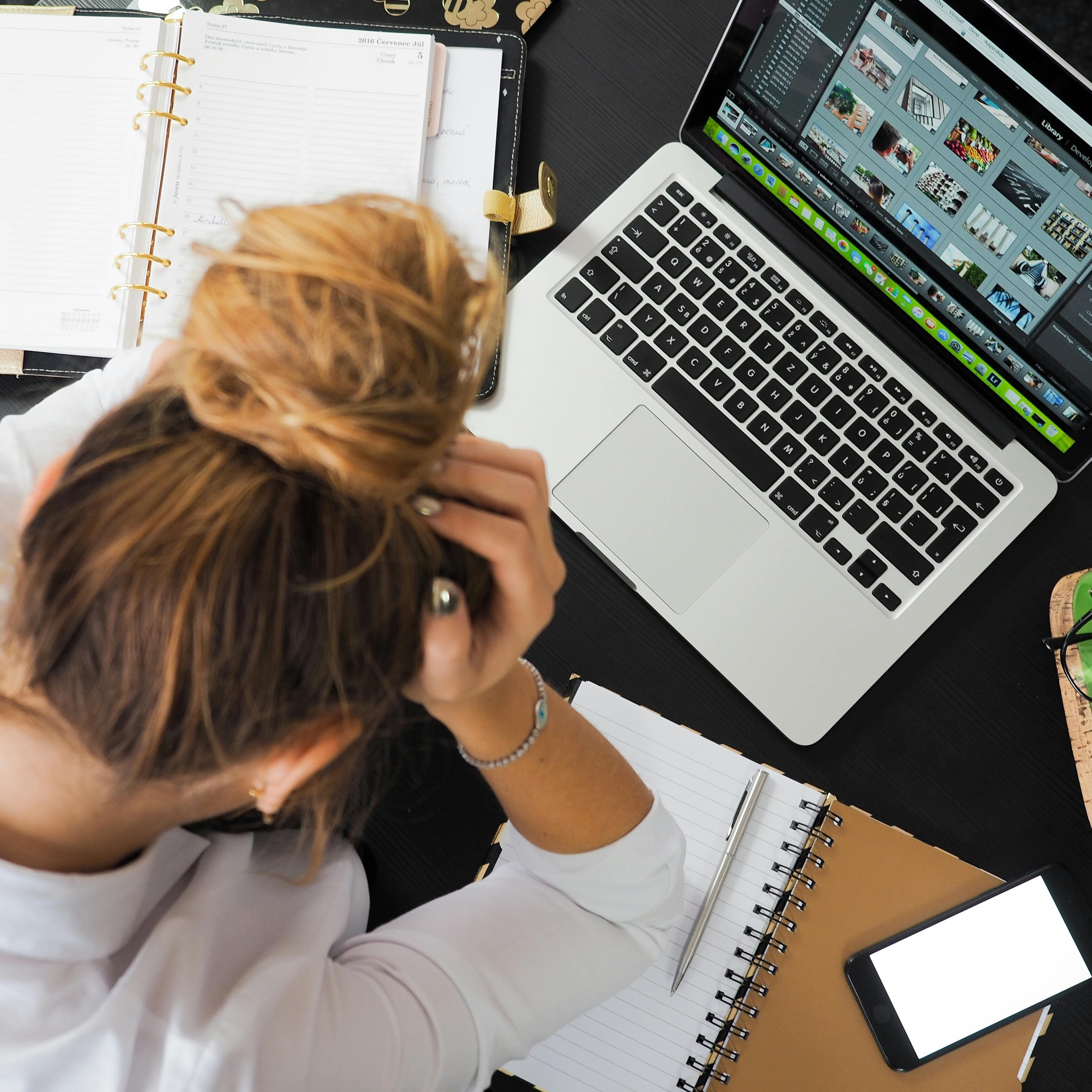 Woman sitting with head in hands, looking down at a computer and scattered notebooks.